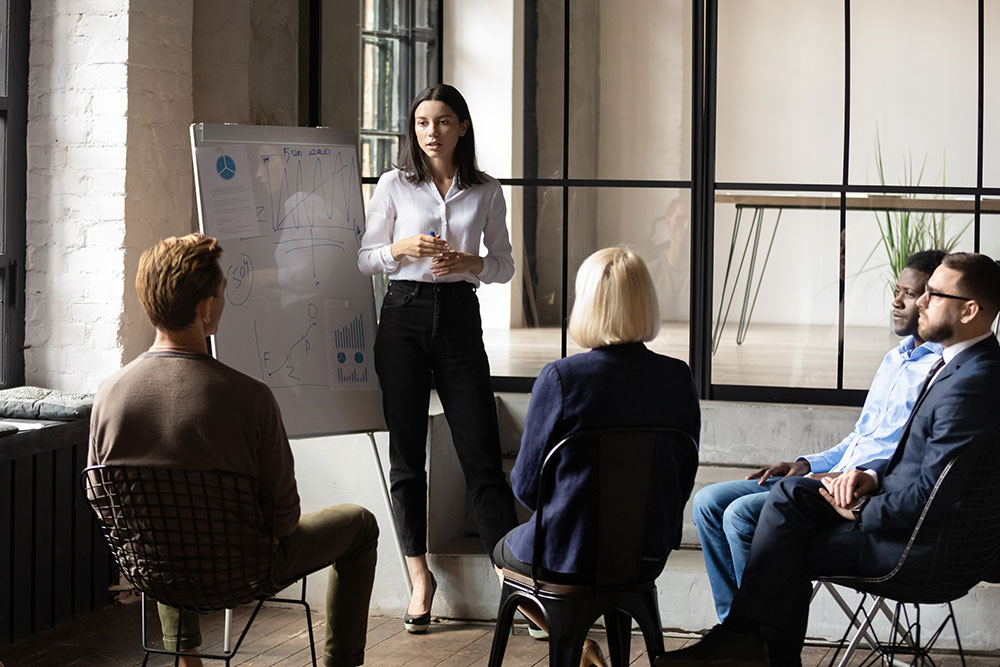 A woman is stood in front of a whiteboard giving a presentation to people in an office.